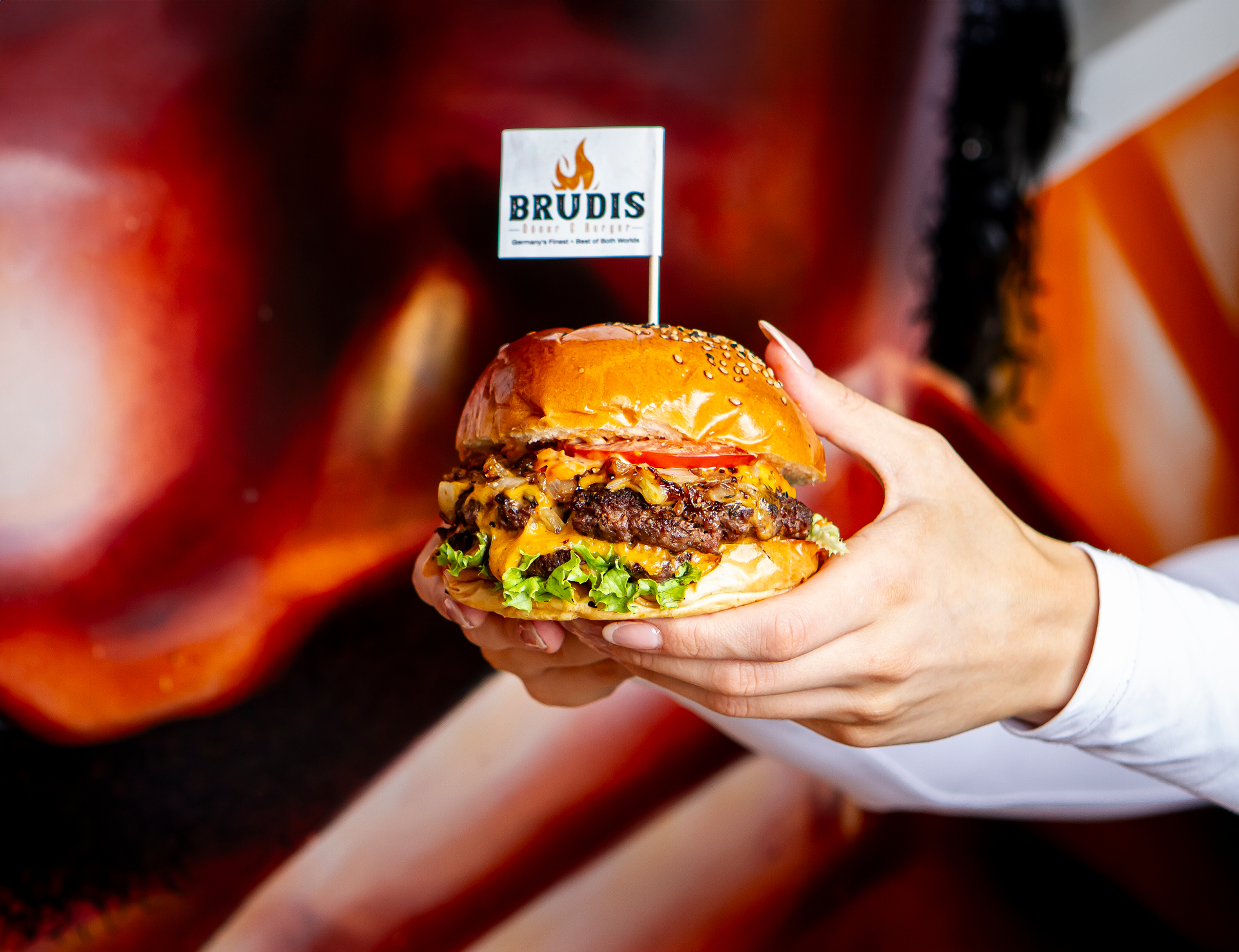 Hands holding a freshly made Brudis smash burger with sesame bun, melted cheese, and Brudis flag